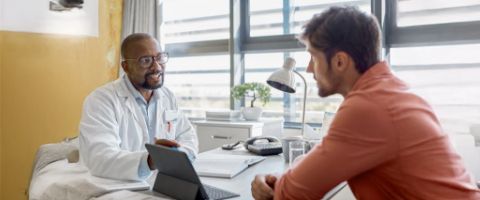 A medical doctor sits at his desk and talks to a patient sitting across from him about information on a tablet propped up on the desk in between them