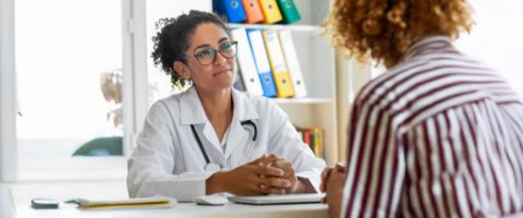 A medical doctor sits at her desk, listening to a patient who is sitting across from her