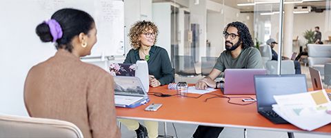 Three people sit at a shared desk with laptops and have a conversation 