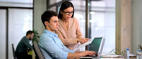 Two people in business casual attire looking at a laptop and paper in an office setting.