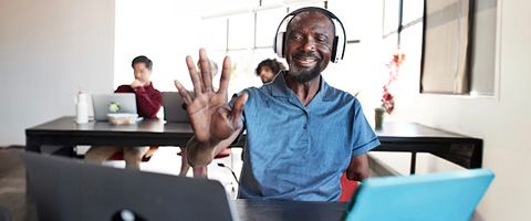 Man in a blue shirt and headphones sitting at a desk while waving at one of two laptop devices in front of him