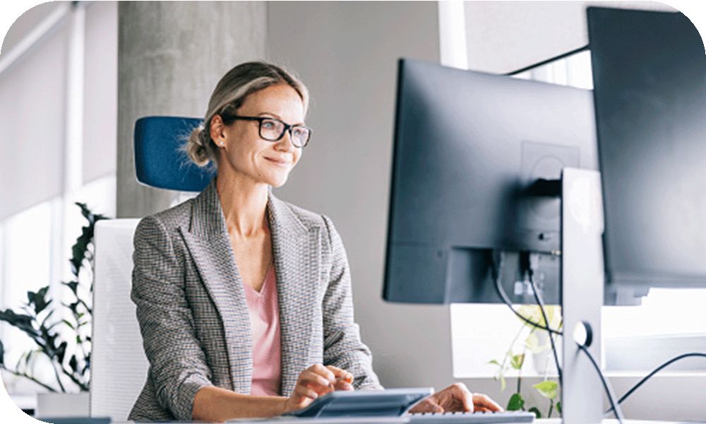 Woman in a grey suit jacket and glasses sitting at a desk while working on a desktop monitor in front of her