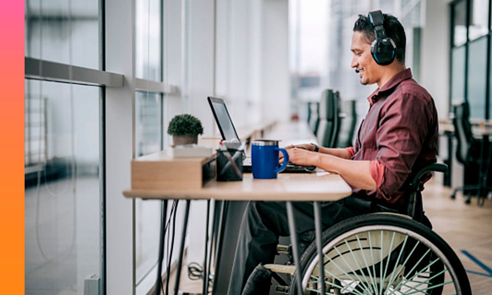 Man in a red shirt sitting in a wheelchair with headphones on while working on a laptop device in front of him