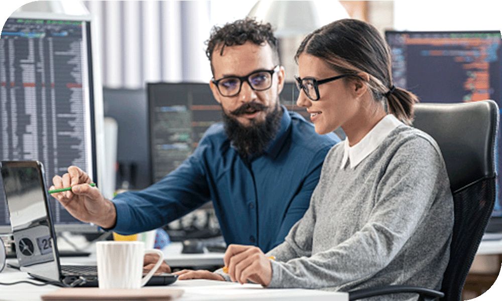 Woman in a grey sweater sitting with a man in a blue shirt in an area filled with computer monitors while working on a laptop device in front of them