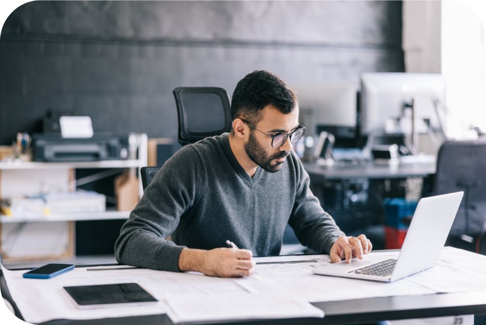 Man in a gray sweater sitting at a desk while working on a laptop device and taking notes