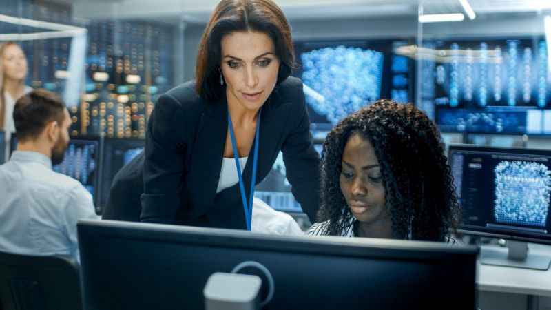 Two female IT professionals working together at a desktop computer in a network operations center.