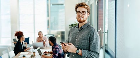 A person holds a phone and smiles at the camera in front of a table with laptops and people seated in a bright office.
