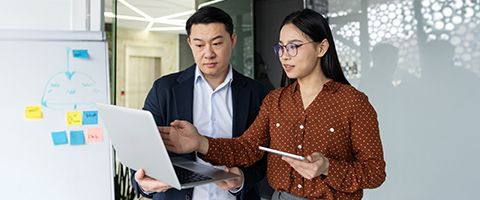 A person holds a laptop while another gestures toward it while holding a mobile phone.