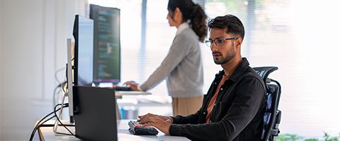 A seated person works at a computer while a standing person works at a different computer in the background.