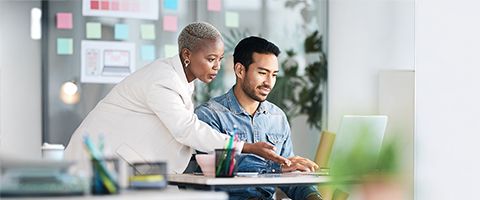 A person leans over a desk to point at a laptop that another person is working on.