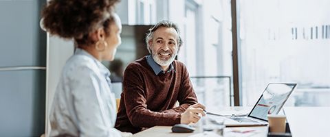 Two people smile and talk while seated at a desk in a bright office.