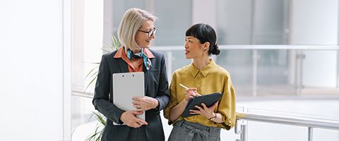 Two businesspeople carry clipboards and talk in a bright office.