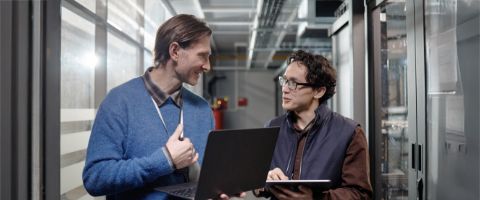 Two people are in conversation in a professional setting, with one holding a laptop and one holding a tablet.
