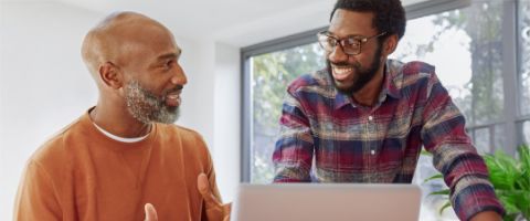 Two people, one standing and one sitting, smile at each other with a laptop in front of them. 