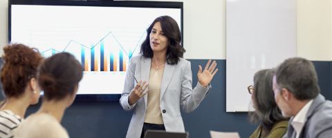 A person in a gray jacket gives a presentation in front of a projector screen showing a graph.
