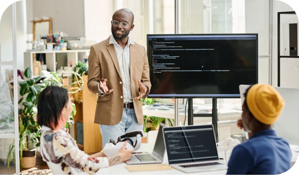 A person in a tan jacket talks to two people who are seated at a table with computers in a bright office