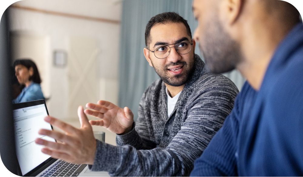 A person with glasses talks to another person sitting next to them in front of a laptop. 