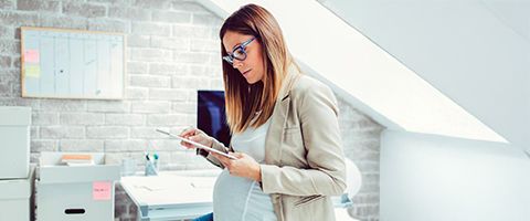 A pregnant person stands in their office while working from a tablet. 