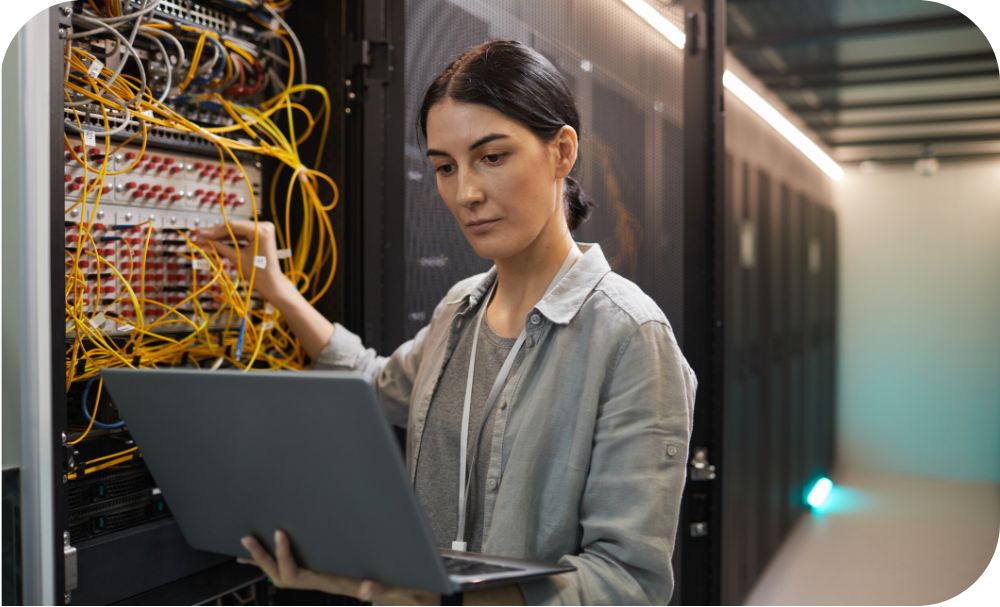 An engineer dressed in business casual holds a laptop as they fidget with the wiring of a server stack beside them. 