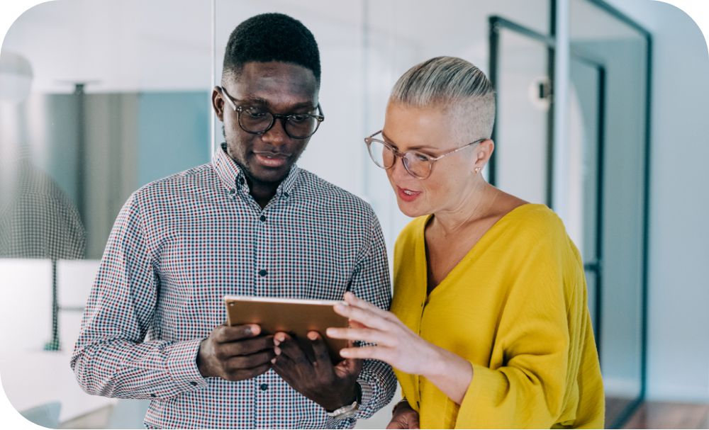 Two professionals in a modern office space stand close together as one leans over the others shoulder to look at a tablet held out between the two. 