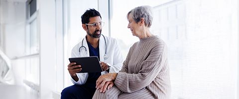 A doctor holding a tablet device and a patient sit and talk near a bright window.