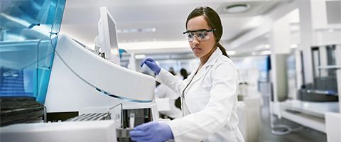 A lab researcher in a lab coat and goggles uses a machine.