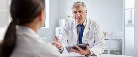 A doctor holds a tablet device and speaks to a patient in a large exam room.