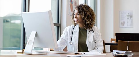 A doctor works at a desktop computer near a window.