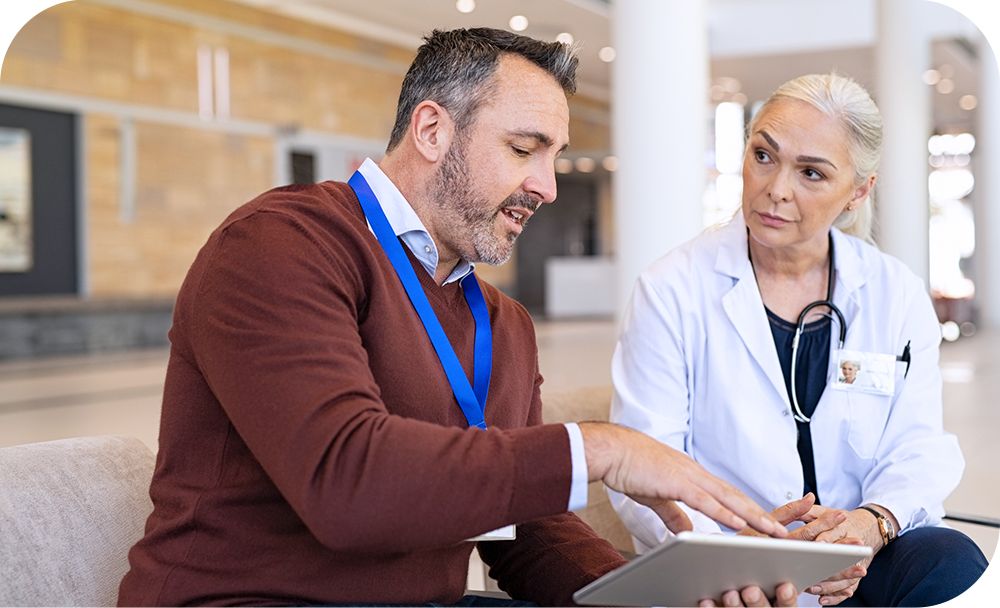 A doctor and a person in casual business attire sit on a bench in a large building and talk over a tablet.