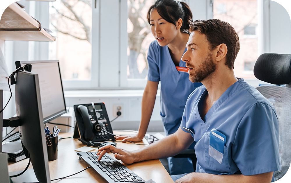Two healthcare professionals wearing blue scrubs working at a desk, reviewing information on a computer monitor in an office setting with medical equipment nearby.
