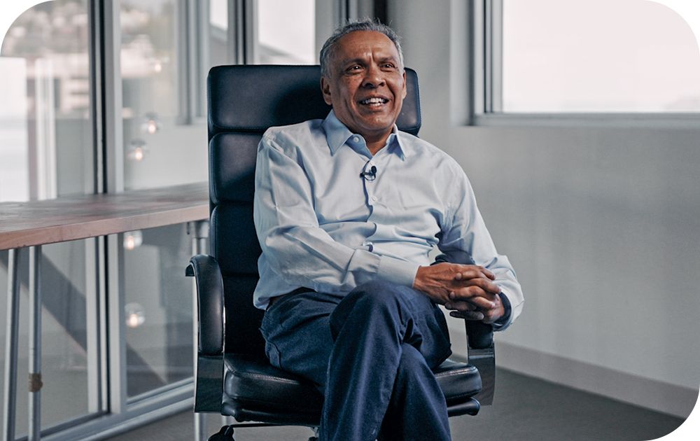 A person wearing a light blue button-down shirt and dark pants smiles while sitting in a black leather office chair in front of a large window.