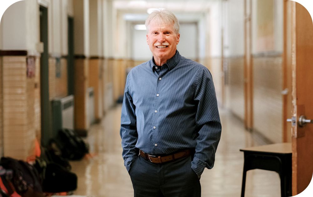 Business professional dressed in a navy, dotted button-down and black slacks, stands smiling with their hands in their pockets before a faded school background.