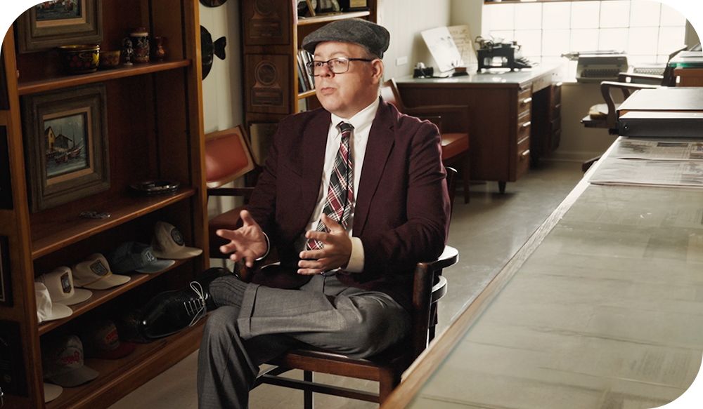 Person in formal clothing sits in an cozy office setting while speaking with hand gestures.
