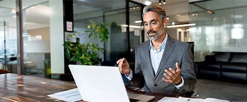 Person in business attire sitting at a conference table in an office setting talking on a laptop.