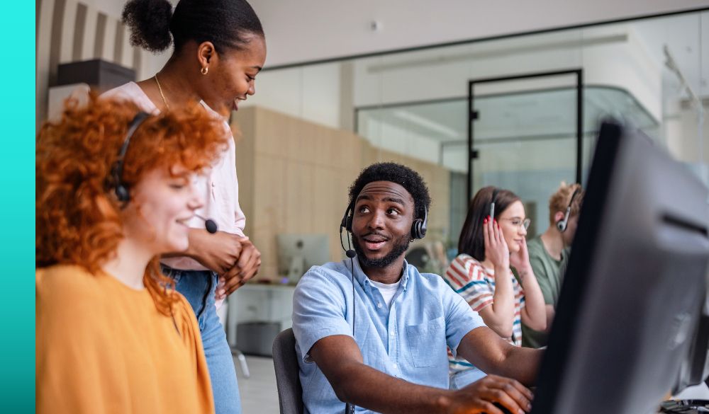 Diverse contact center employees wearing headsets engage in conversation while working at computers.