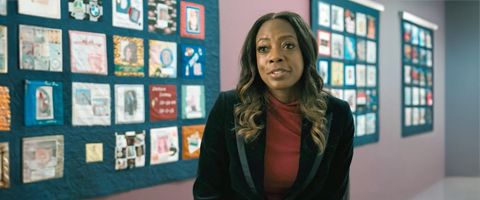 Businessperson in a black blazer and maroon top speaks in front of a wall-mounted quilt featuring various images. 
