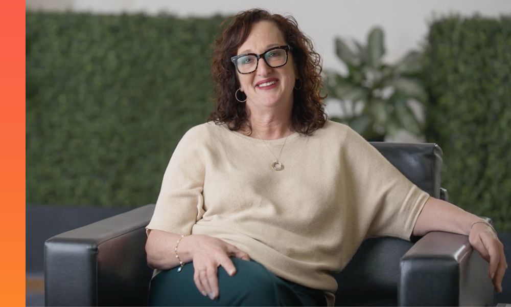 Businessperson in a black blazer and maroon top speaks in front of a wall-mounted quilt featuring various images. 