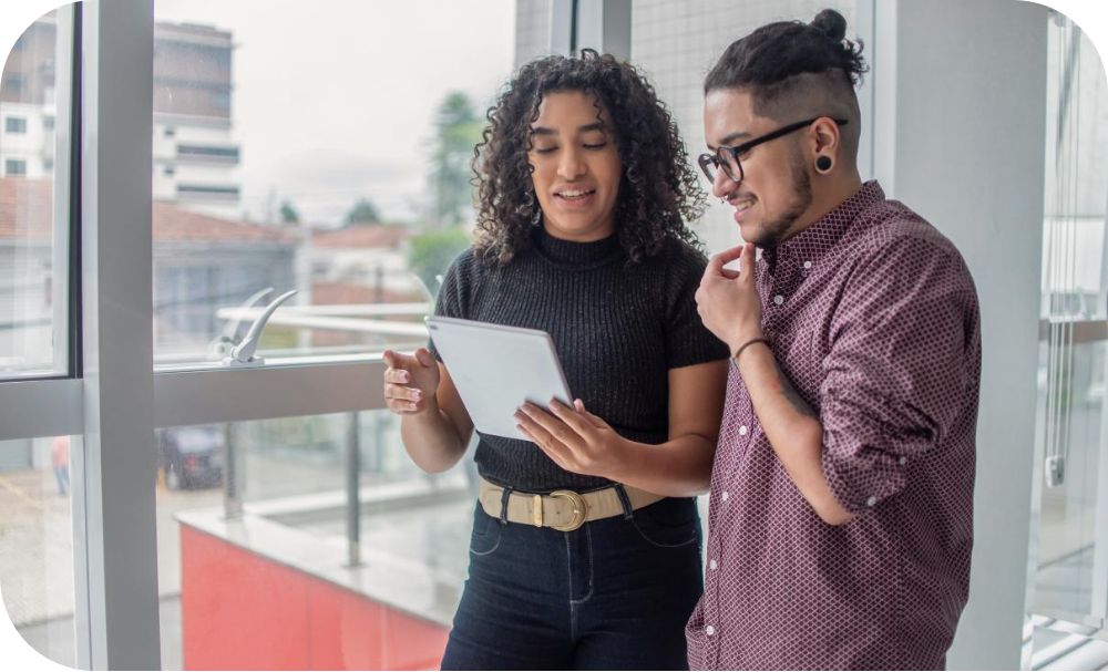 Two individuals stand next to a window and look over a tablet one of them is holding.