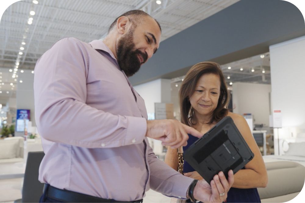 Two individuals in a furniture store stand by each other and look at a tablet one of them holds.