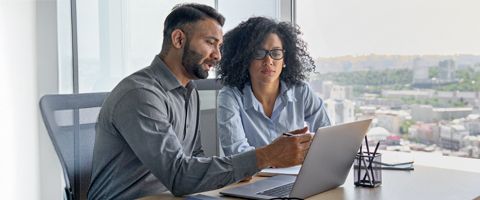 Two businesspeople sit at a desk and collaborate while referencing something on a laptop screen. 