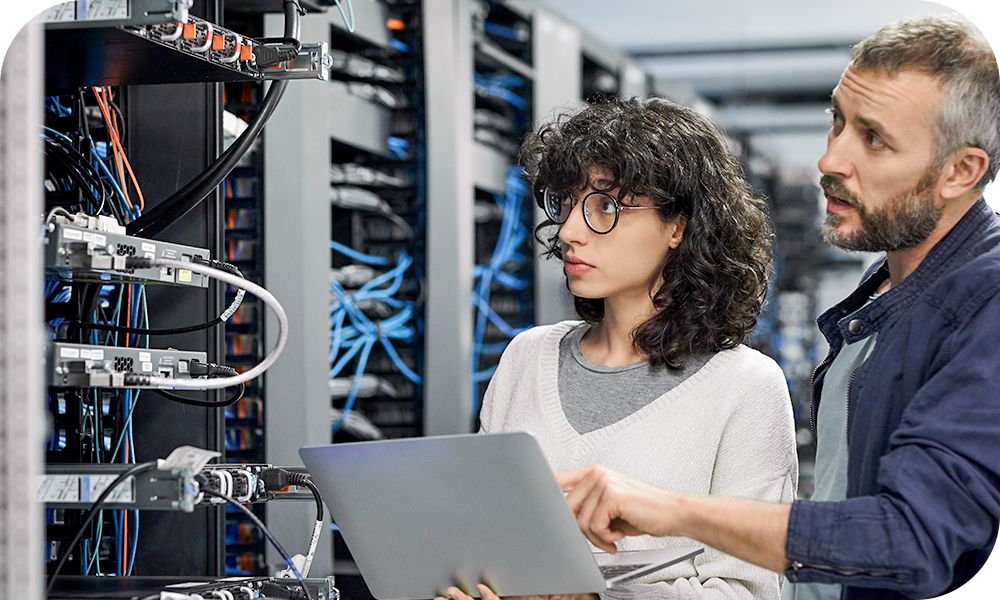 Two professionals look at a server stack, with one holding a laptop and the other pointing at it as they start to speak. 