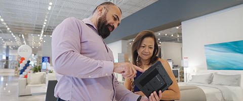 Two individuals in a furniture store stand by each other and look at a tablet one of them holds. 
