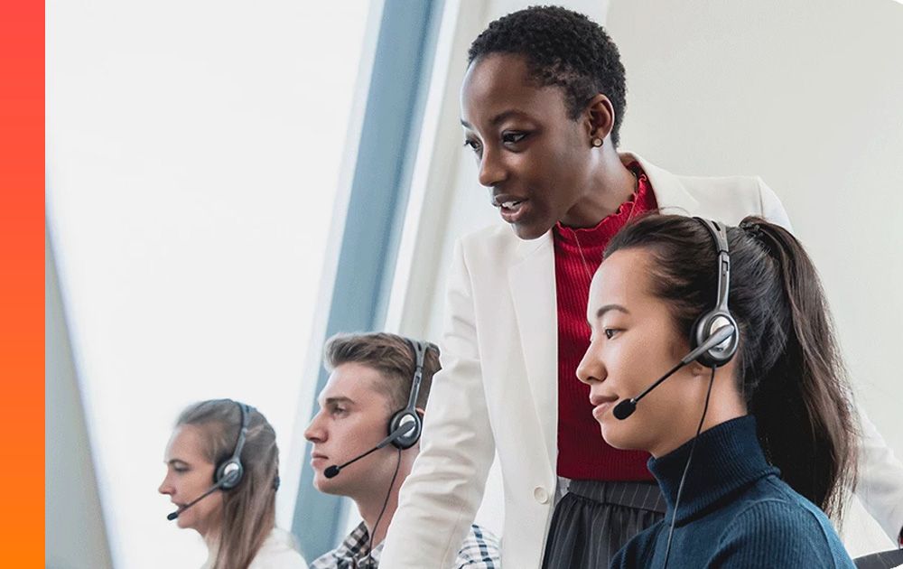Three people with headsets sit and work while another person stands between two of them.