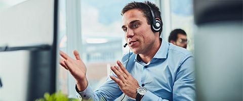 A person in a blue button-down shirt talks on a headset near a window.