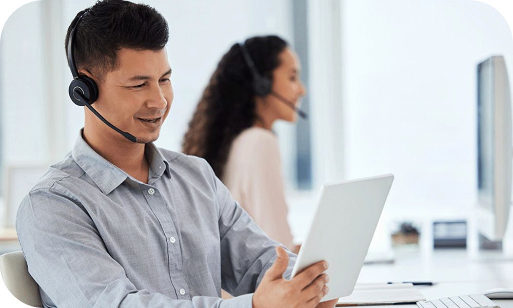 A person wearing a headset works on a tablet while another person works in the background in a bright office.
