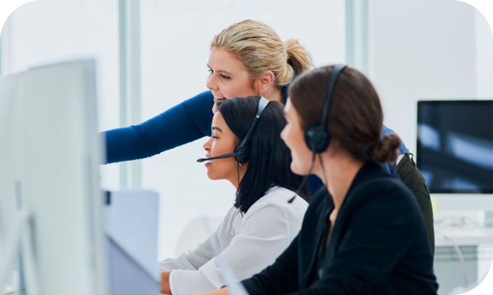 Two call center workers wearing headsets sit at their computers while a person standing next to them points at something on the screen.