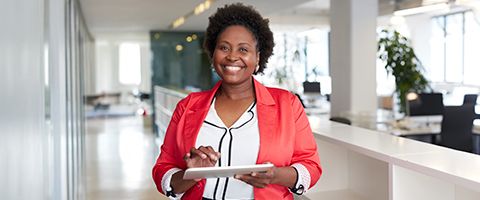 A person in business casual attire smiling and holding a tablet in an office setting.