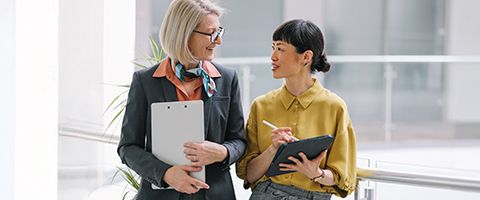 Two people in business casual attire holding tablets and talking in an office setting.