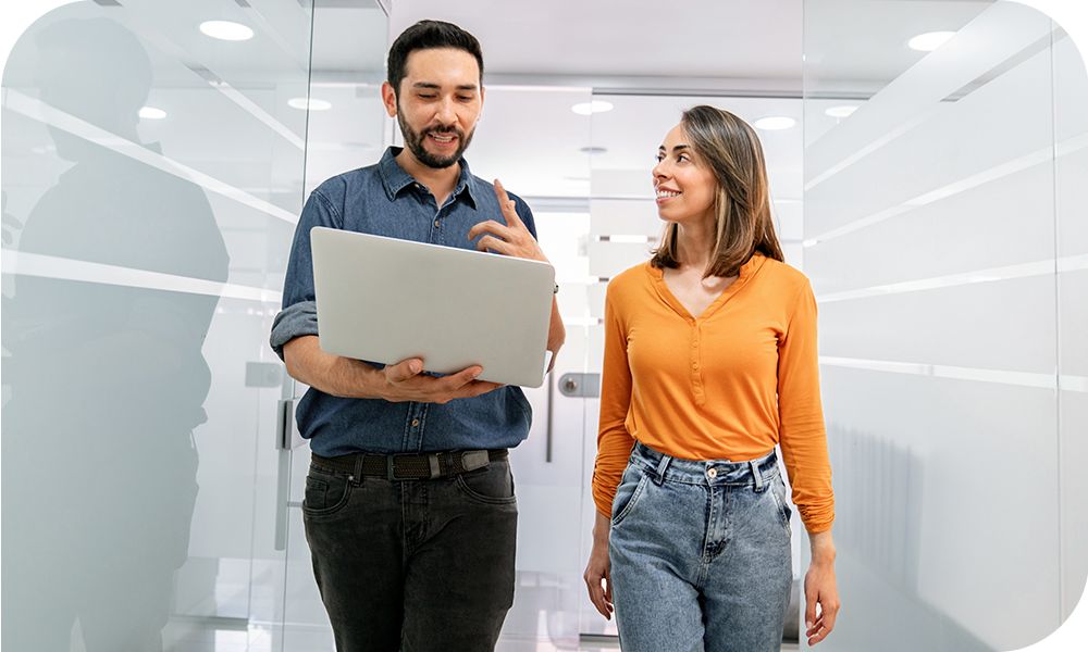 Two people in casual attire walking down a white hallway talking while one holds a laptop.