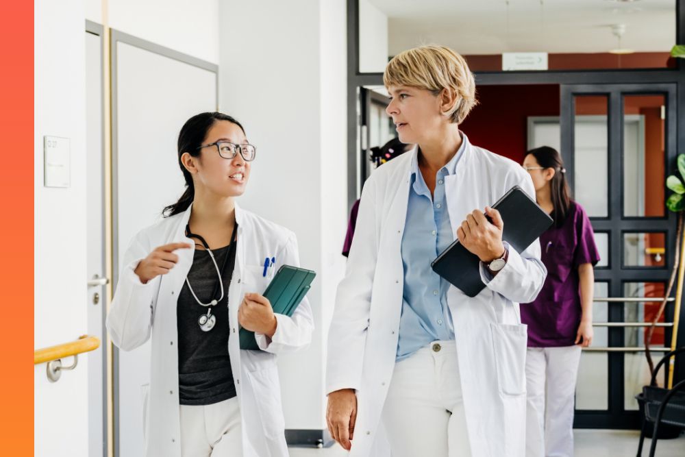 Two women in white doctor's coats walking down a hallway while talking and holding charts 